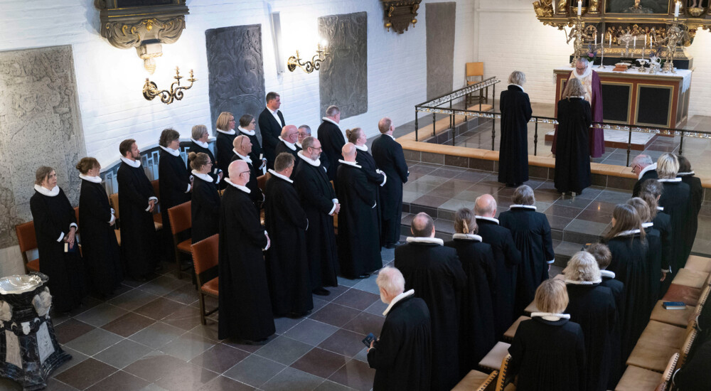 En flok præster står op og kigger på de to præster ved alteret foran biskoppen